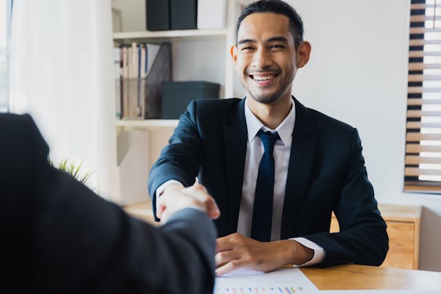 Um homem asiático vestindo terno sorrindo e apertando as mãos de alguém do outro lado da mesa.